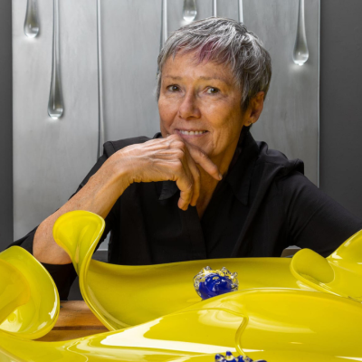 A person sits behind yellow glass art pieces, smiling with hand on chin.