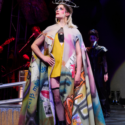 Person in colorful, graffiti-style cape poses on stage with musicians and dramatic lighting.