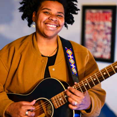 Person smiling and playing a black acoustic guitar indoors, wearing a brown jacket.