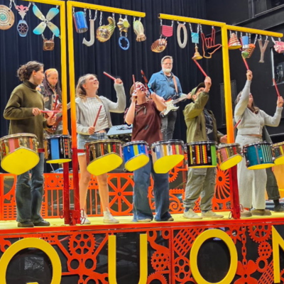 A band performs on a colorful stage with drums and hanging objects, SQUONK displayed below.