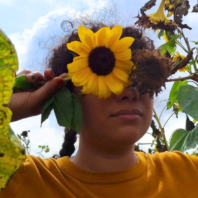 Person holding a sunflower in front of their face in a field, wearing a yellow shirt.