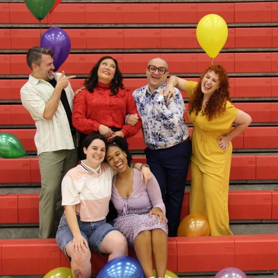 Six smiling adults pose together on red bleachers with colorful balloons. Two people sit in front, while four stand behind them. They appear happy and playful in casual and bright clothing.