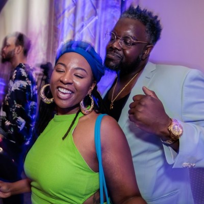 Two people smile and pose for the camera at a lively indoor party, with purple and blue lighting. Other guests are dancing and mingling in the background. The woman wears a green dress and blue hat, and the man wears a light blue suit.
