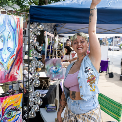 Smiling person raises arm at outdoor art booth with colorful abstract paintings and decorations.