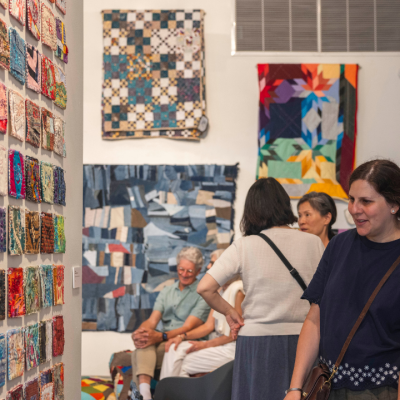 People observe colorful quilts and fabric squares displayed on gallery walls.