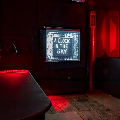 A dimly lit room with dark walls illuminated by red lights. A bulletin board with papers is on the left, and a screen at the back displays the text A LOCK IN THE SKY. Wooden floors and benches are visible
