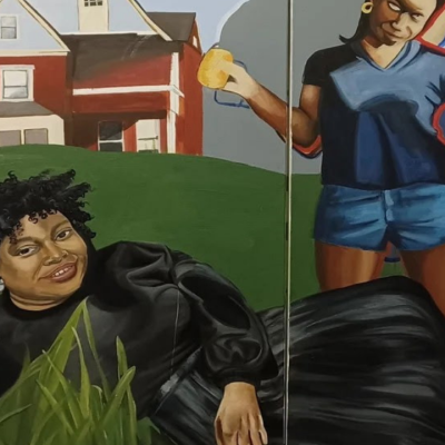 Two women pose on grass; one reclines, one stands, with a red house in the background.