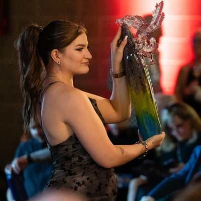 A woman in a formal dress holds up a colorful glass sculpture on stage in front of an audience, with red lighting in the background.