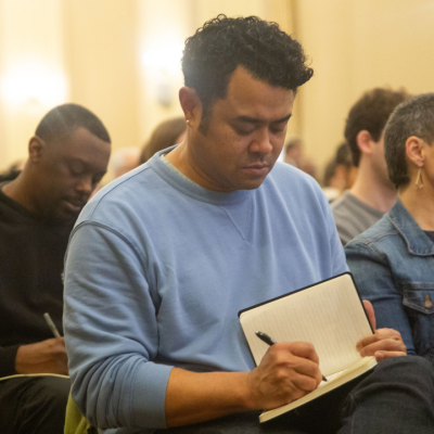 People sitting in an audience, writing in notebooks and listening attentively.