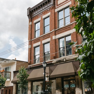 A three-story brick building with large windows and brown awnings over the ground-floor shops, situated on a city street with trees and a lamp post in front.