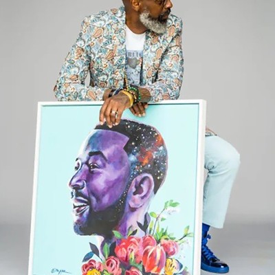 A bald Black bearded man wearing glasses, a white t-shirt, a patterned blazer, light slacks, and blue-and-black shoes. He's sitting down and posed in front of a large framed illustration of a man with flowers