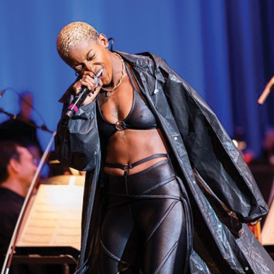 A Black musician with a blonde buzz cut sings into a microphone. They're wearing a black bra top, black pants, and a long black jacket. Behind them are concert musicians and a blue stage curtain