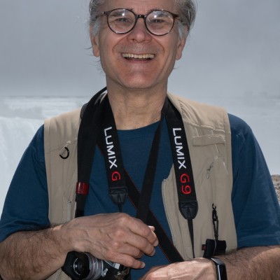 A smiling white man with gray hair wearing black glasses, a blue t-shirt, a tan vest, and a camera attached to a strap hung around his neck