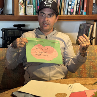 A person holds a handmade sign that reads We commit to equitable artist wages, sitting at a table with craft supplies and bookshelves in the background.