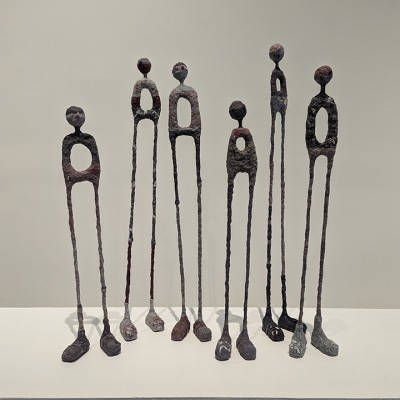 Six skinny sculptures show the rough shapes of human heads and torsos attached to long legs and feet, each with no arms and a hole in each of their stomachs
