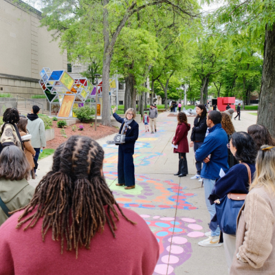 A group of people listen to a guide outdoors near a museum and colorful ground art.