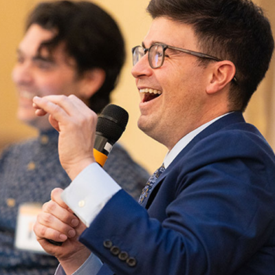 Man in a suit speaking into a microphone while smiling at an event.