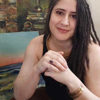 Woman with long brown hair and dreadlocks sits in front of colorful paintings.
