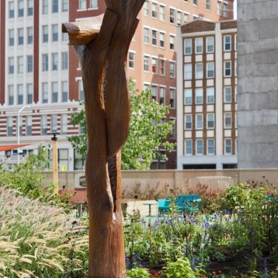 a tall sculpture that looks like it was made out of wood stands on a pedestal in front of city buildings