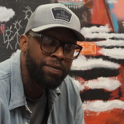 A Black man with a beard, gray baseball cap, black glasses, gray T-shirt, and denim collared shirt poses in front of a colorful painting
