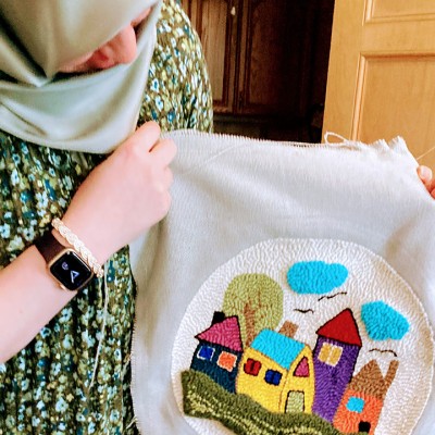 A person holds up a piece of colorful textile artwork of houses and clouds