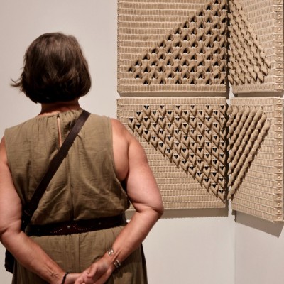 A person in a sleeveless olive dress stands with their hands behind their back, facing a wall-mounted geometric artwork made of textured, folded cardboard or similar material.