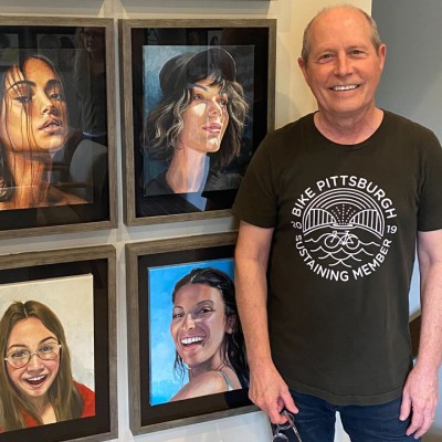 A white man wearing a black Bike Pittsburgh T-shirt and blue jeans stands smiling next to painted portraits hanging framed on a wall
