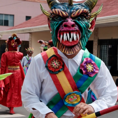 People in colorful demon masks and costumes parade down a street during a festival.
