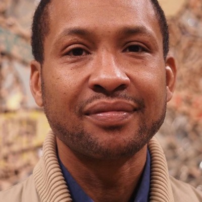 A smiling Black man with short dark hair, a subtle closely-shaved beard, wearing a blue button-down shirt and a tan jacket. Behind him in a colorful tiled artwork