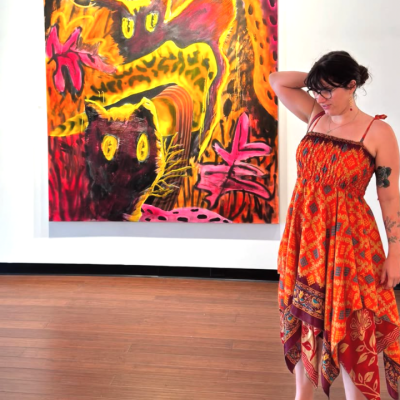 Woman in a red dress stands by a colorful painting of abstract cats in a gallery.
