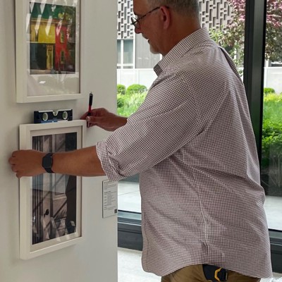 A white man with gray hair and glasses, wearing a button-down dress shirt and a black watch, hangs artwork on a white wall next to a window with green grass and a purple tree