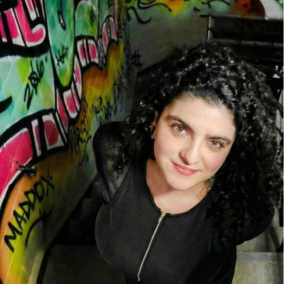 Woman with curly hair stands by graffiti-covered wall, looking up at the camera.