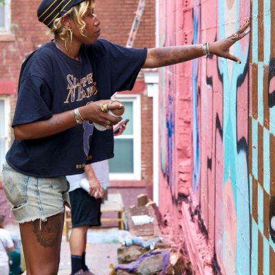 A person in a black hat and t-shirt with denim shorts spray paints a colorful mural on a brick wall. Another person stands nearby. The scene is outdoors, between buildings, with mural supplies visible.