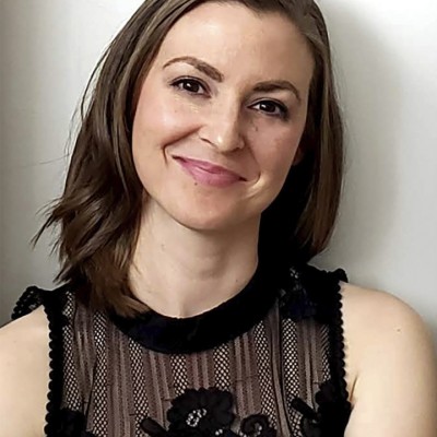 A smiling white woman with shoulder-length straight brown hair, wearing a sleeveless black shirt