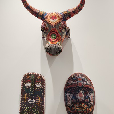 Three Mexican masks hanging on a wall, depicting an antlered animal on top and two faces on the bottom