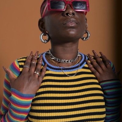 Black person with short hair, red sunglasses, large silver hoop earrings, silver necklaces, and a colorful striped long sleeved shirt