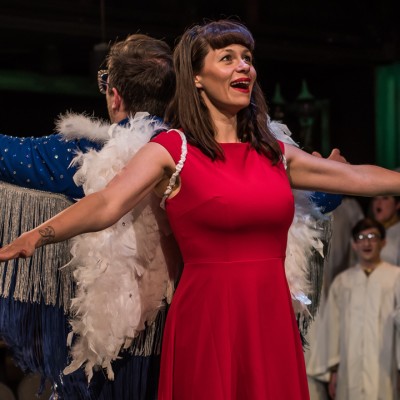 Two actors stand back-to-back on stage. One is a white woman with long brown hair and brown bangs, wearing a red short-sleeved dress, and white angel wings strapped to her arms. Behind her stands what looks like a white man with short brown hair, wearing a blue jacket