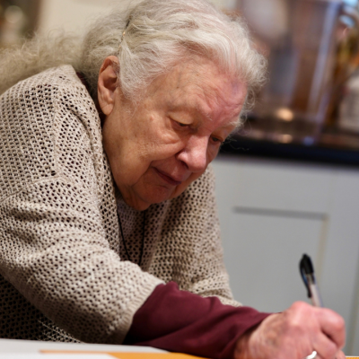 An older woman with white hair writing on paper with a pen.