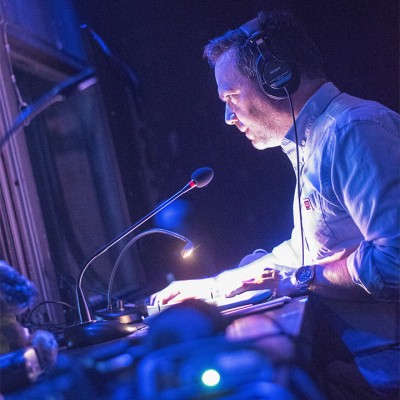 Photo of Nathan Ruggles. Inside a theater tech booth, from a vantage point low above a long desk running away along its length, he sits in profile, leaning toward a mic sitting on the cluttered surface, gazing intently out through the window at left. A middle-aged male wearing headphones and a sky blue oxford cuffed at the elbow, a small table lamp reflects striking white light up into the light pink skin of his face, casting deep shadows in what otherwise is a space dimly lit in blue and lavender.