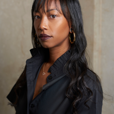 Woman with long dark hair and black shirt standing against a beige background.