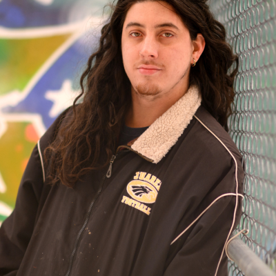 A person with long hair leans against a chain link fence outdoors.