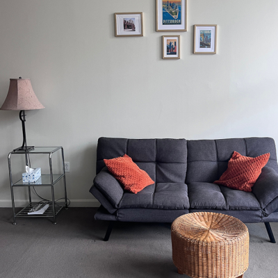 A living room with a gray sofa, two orange pillows, a round wicker coffee table, a metal side table with a lamp and tissue box, and five framed pictures on a plain white wall.