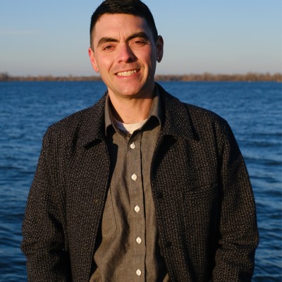 A white man with short dark hair smiles while standing in front of a body of water