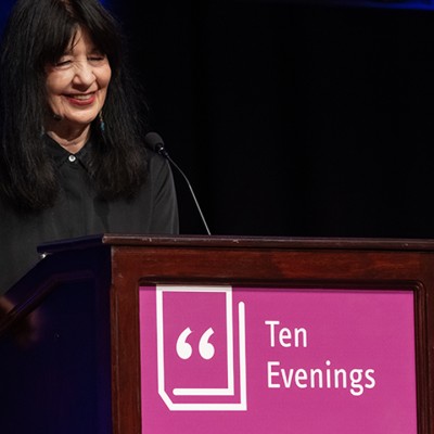 A woman with dark hair stands smiling behind a podium that reads, "Ten Evenings, Pittsburgh Arts & Lectures"