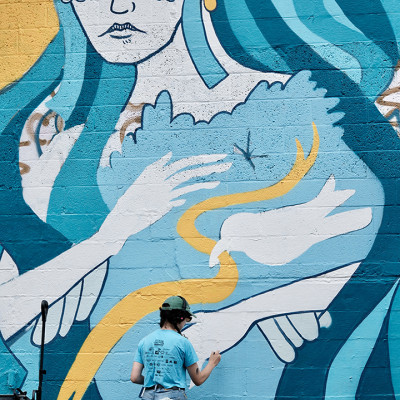 A person in a blue shirt and helmet paints a large mural of a woman with blue hair holding a white bird on a brick wall. The mural features bold lines and shades of blue, yellow, and white.