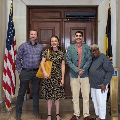 A group of four people stand in a government building, in front of large wooden doors and in between the American Flag and the City of Pittsburgh Flag. 