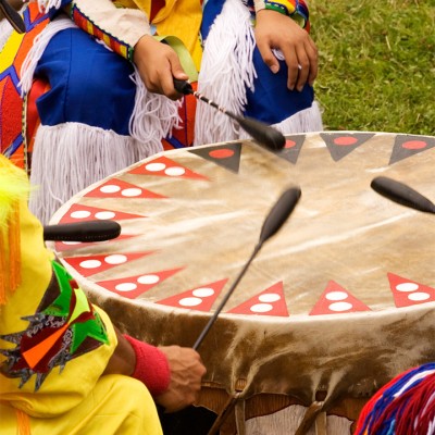 Native Americans in colorful outfits sit in a circle around a drum