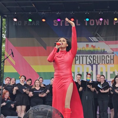 Person wearing a long red dress sings into a mic while holding one arm in the air. A choir of people dressed in black clothing stand behind them on stage in front of a rainbow flag