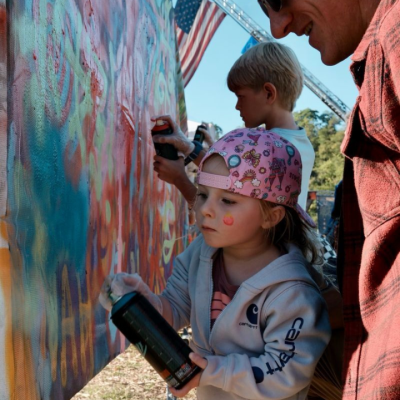A young child in a pink cap spray paints a colorful mural while a man watches.