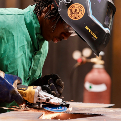 A person wearing safety gear and a welding helmet uses a power grinder on a metal surface, creating sparks. A gas cylinder is visible in the background. The helmet has a Weld For Community sticker.